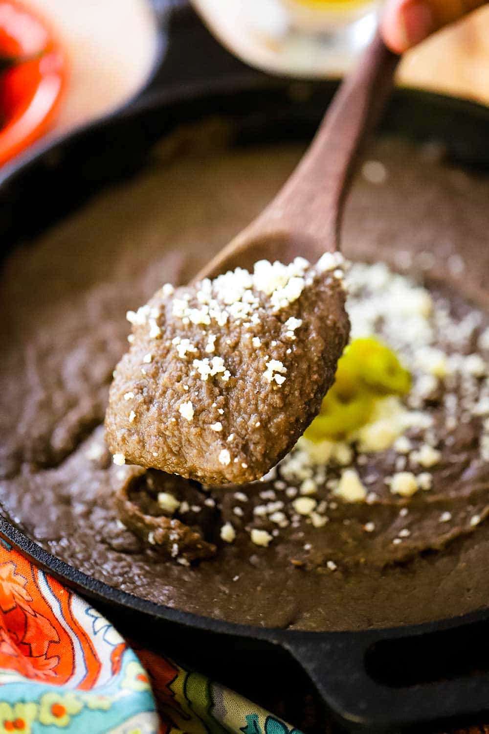 A large wooden spoon full of refried beans topped with crumbled Cojita cheese being lifted from a pot of the beans. 