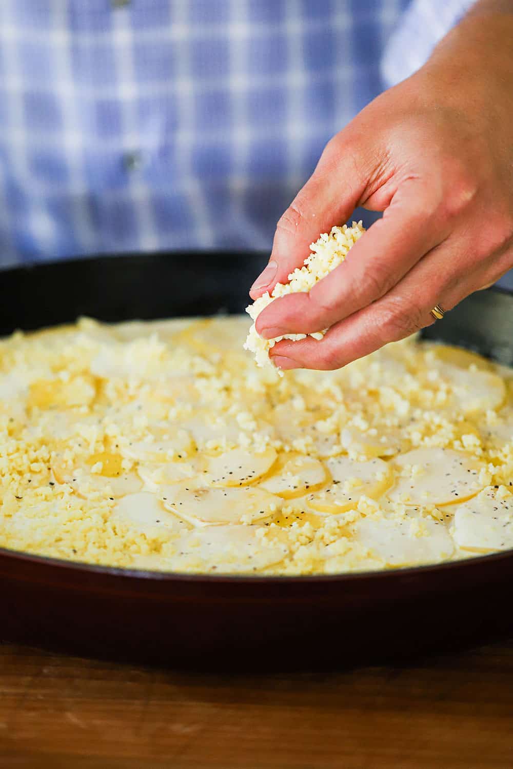 A person sprinkling shredded Gruyere cheese over layers of potatoes in cream all in a oval baking dish.