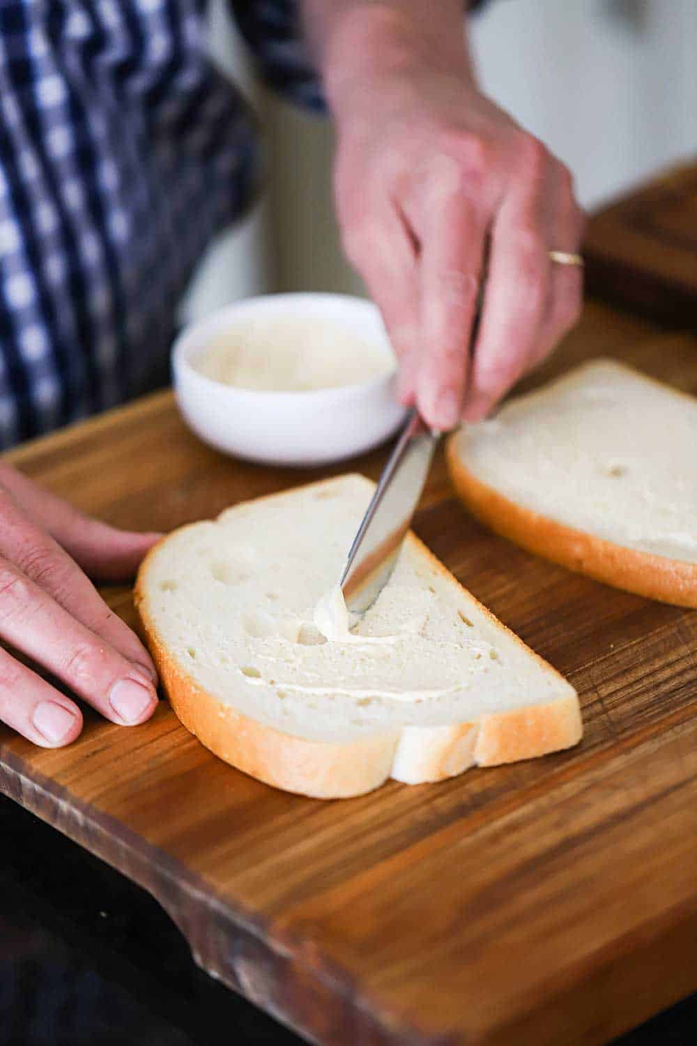 A person using a knife to spread a mayonnaise and Dijon mustard mixture on one side of sliced white bread. 