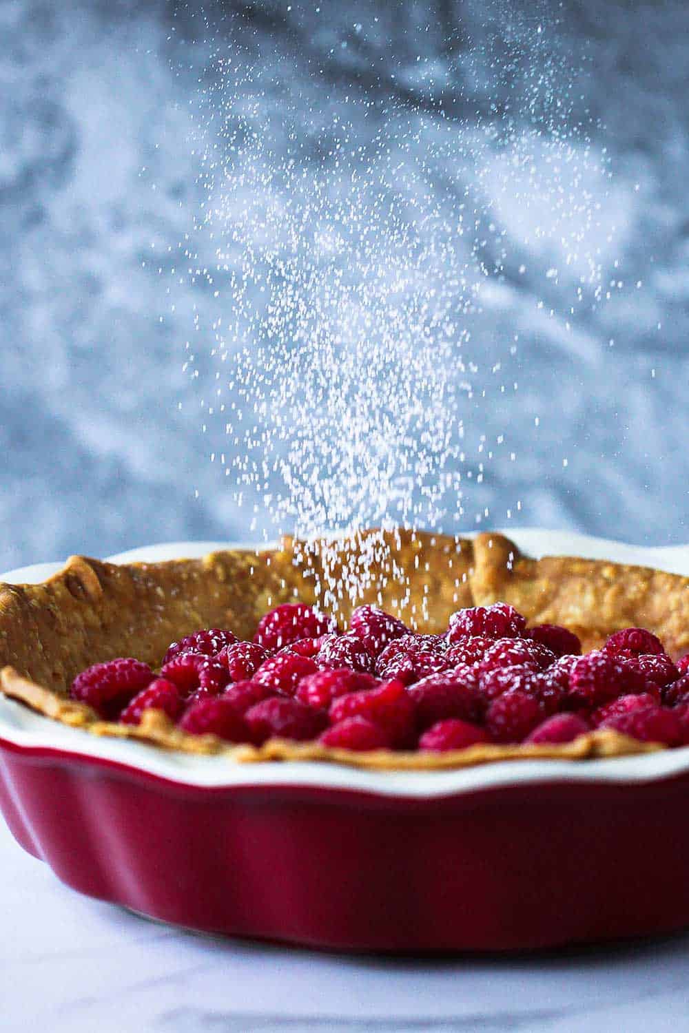 A chess pie in a red pie dish topped with fresh raspberries being sprinkled with powdered sugar from above.