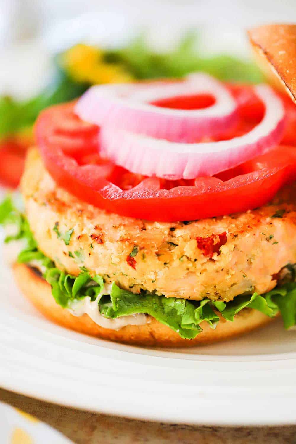 A fully cooked salmon burger on a hamburger bun, garnished with lettuce, tomato, and red onion all sitting on a white plate.