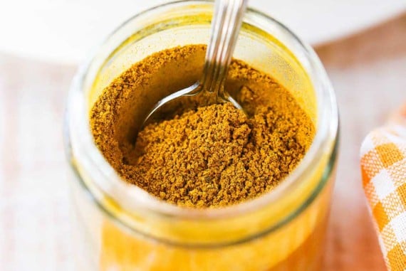 A small glass jar that is filled with Jamaican curry powder with a spoon inserted into it, and sitting next to an orange checkered cloth.