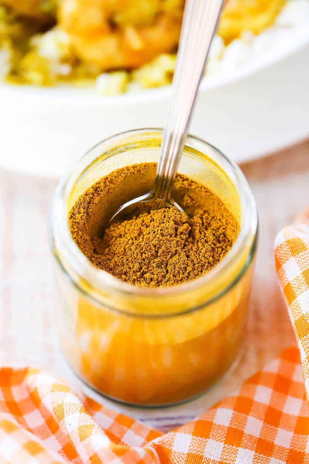 A small glass jar that is filled with Jamaican curry powder with a spoon inserted into it, and sitting next to an orange checkered cloth. 