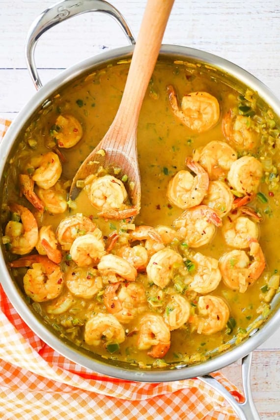A large stainless steel skillet filled with shrimp curry in a sauce with a wooden spoon insert into the middle of it.