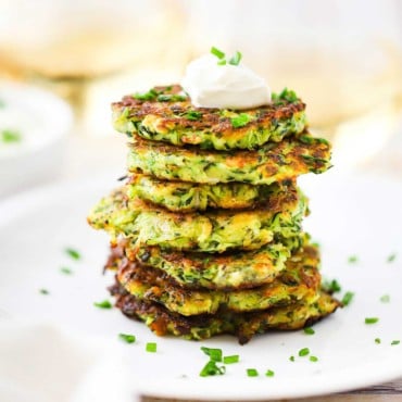 A stack of zucchini fritters on a white dinner plate topped with a dollop of sour cream and snipped chives sprinkled all over.