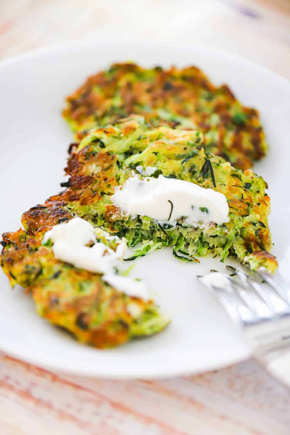 A zucchini fritter that has been split open with a fork and topped with a dollop of sour cream on a white dinner plate. 
