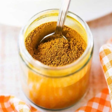 A small glass jar that is filled with Jamaican curry powder with a spoon inserted into it, and sitting next to an orange checkered cloth.