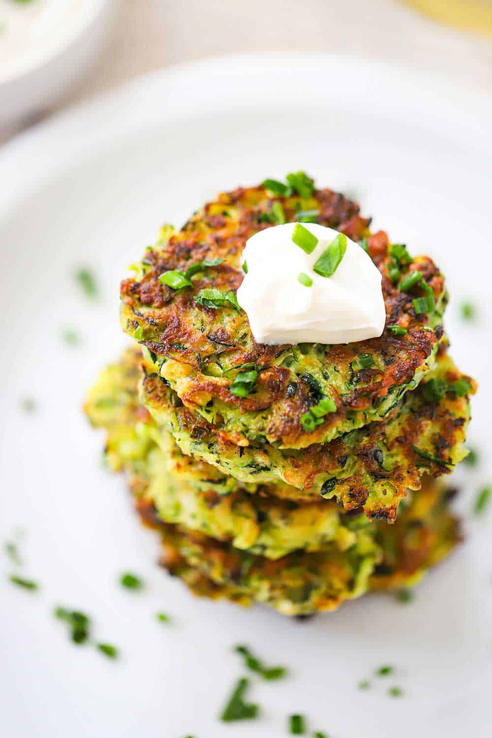 A stack of zucchini fritters on a white plate with a dollop of sour cream on the top fritter and sprinkled with snipped chives. 