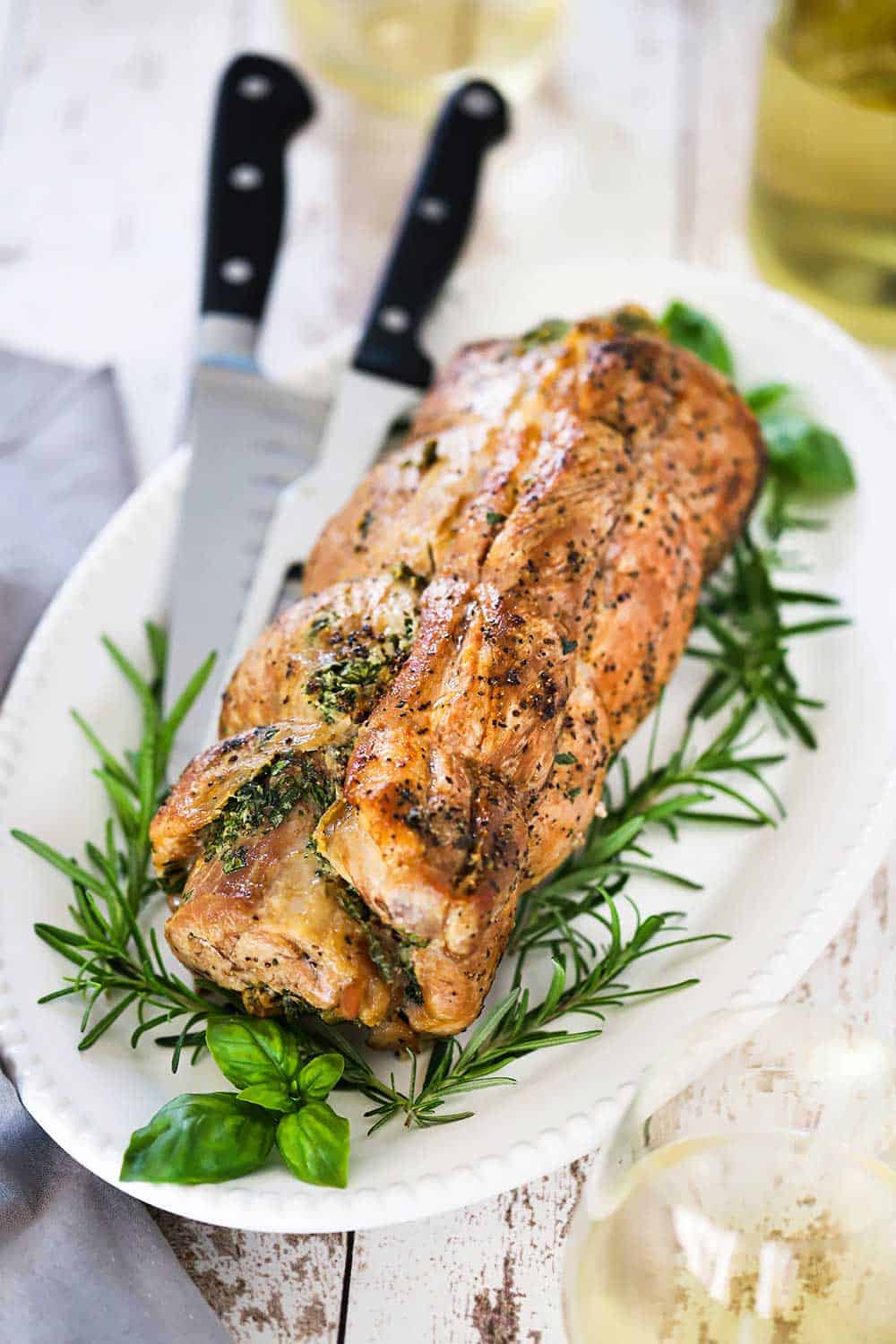 An herb-stuffed pork loin roast sitting on an oval platter with a large knife and fresh herbs nestled around it.