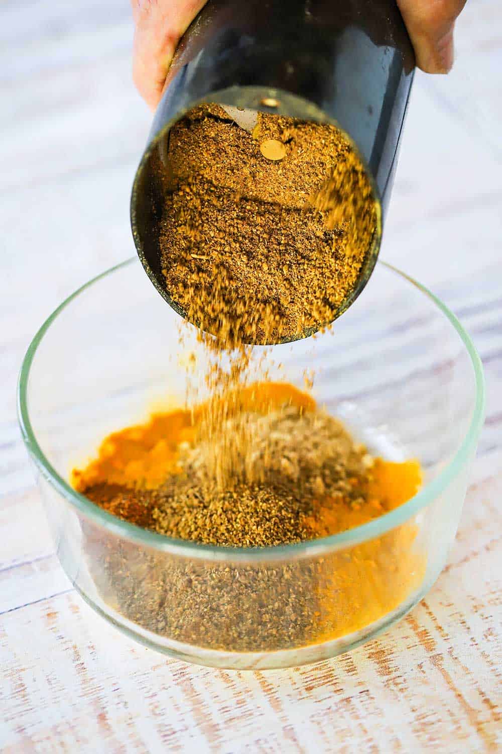 A person pouring ground toasted seeds into a glass bowl filled with ground turmeric and other ground spices. 