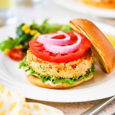 A fully cooked salmon burger on a hamburger bun, garnished with lettuce, tomato, and red onion all sitting on a white plate.