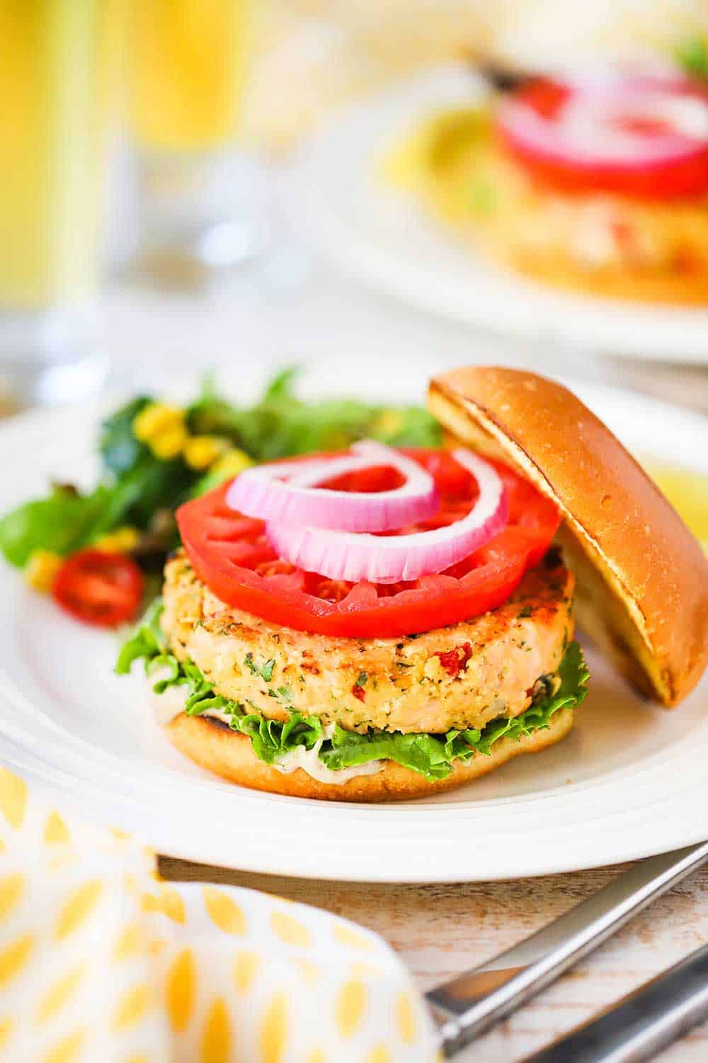 A fully cooked salmon burger on a hamburger bun, garnished with lettuce, tomato, and red onion all sitting on a white plate. 