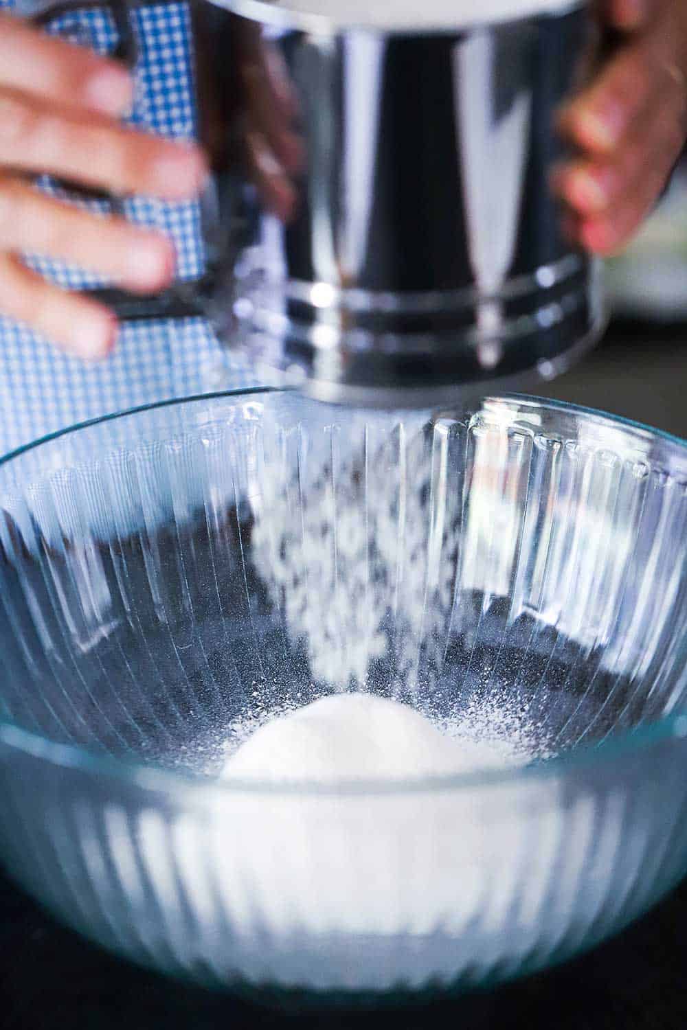 A person holding a metal sifter over a bowl while sifting flour into it. 