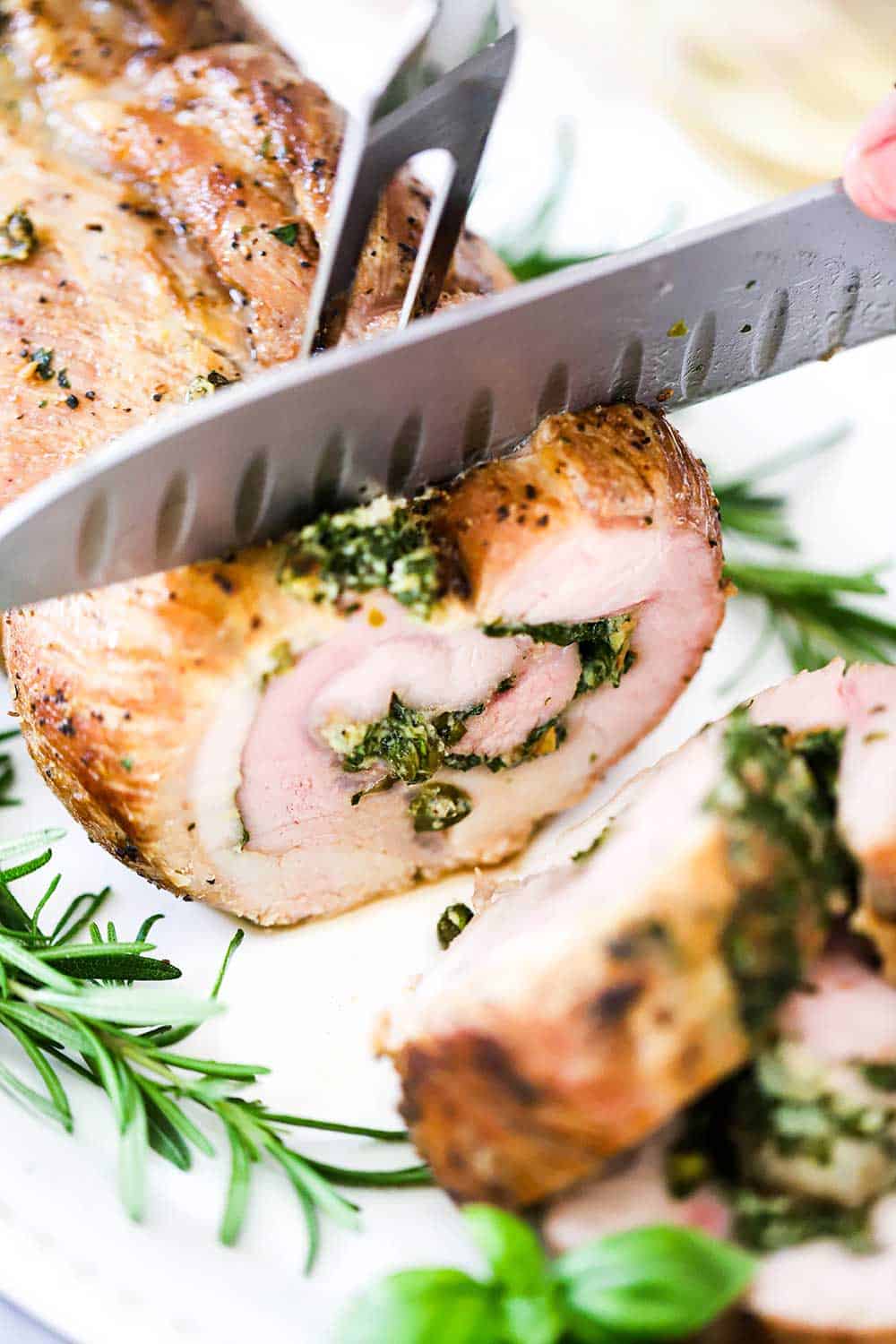 A chef's knife cutting into a herb-stuffed pork loin on a white platter.
