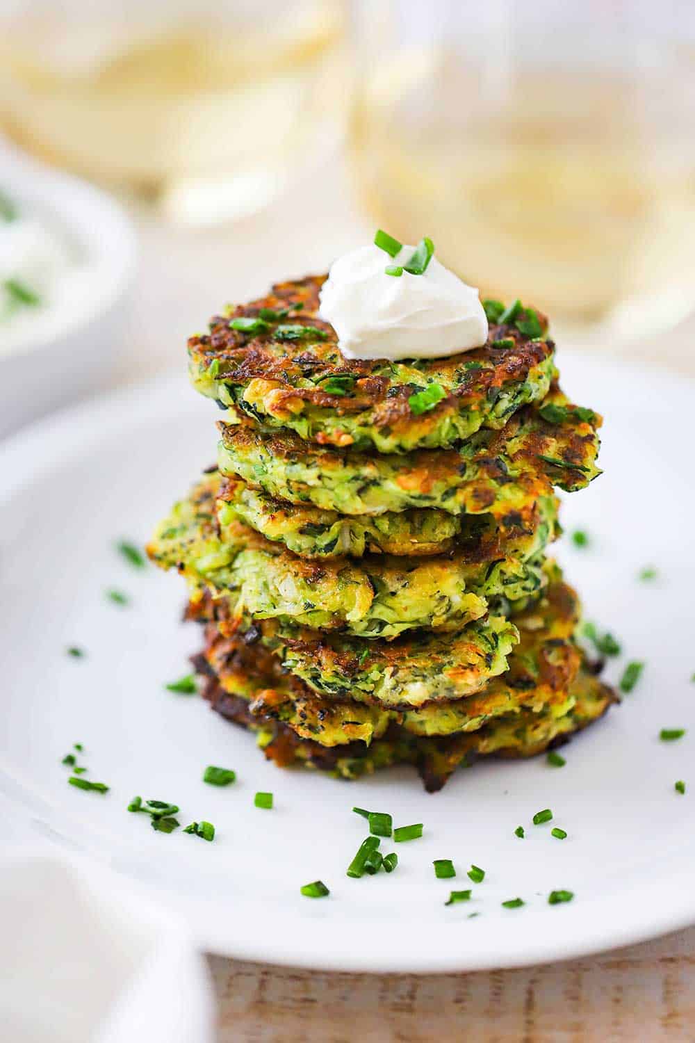 A stack of zucchini fritters on a white dinner plate topped with a dollop of sour cream and snipped chives sprinkled all over. 