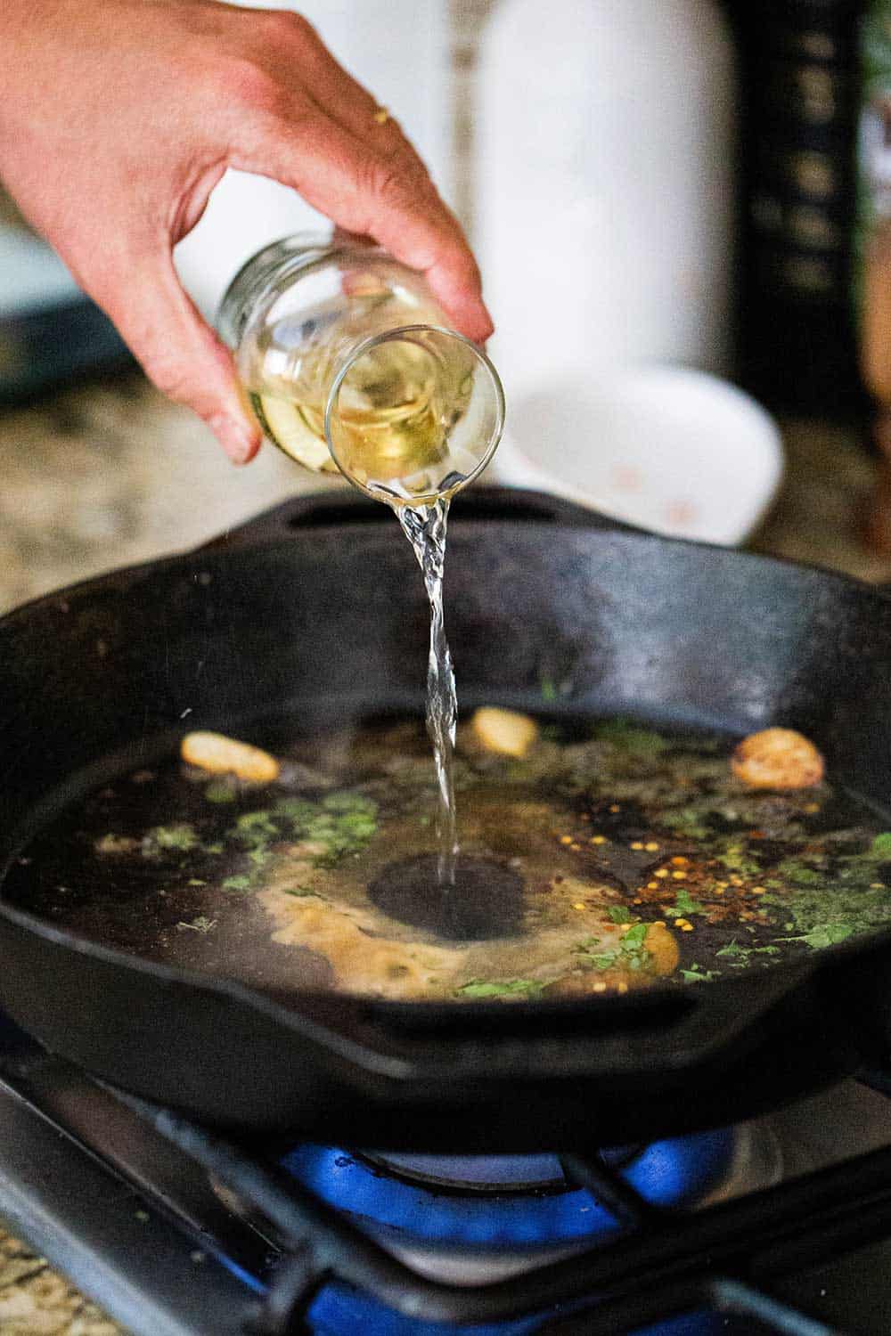 A person pour white wine from a small carafe into a cast-iron skillet will with garlic halves, red pepper flakes, and freshly chopped oregano. 