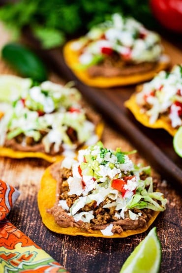 A tostada on a wooden background topped with refried beans, taco meat, lettuce, tomatoes, and Mexican cream, surround by other assembled tostadas.