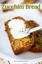 Two slices of zucchini bread being smeared with softened butter all sitting on a white plate.