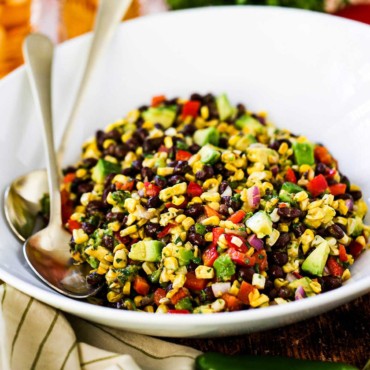 A large white serving bowl filled with black bean and corn salad with two gold serving spoons tucked in beside the salad.