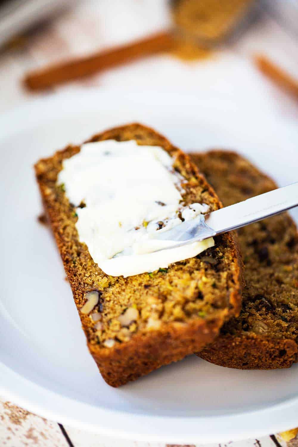 Two slices of zucchini bread being smeared with softened butter all sitting on a white plate. 