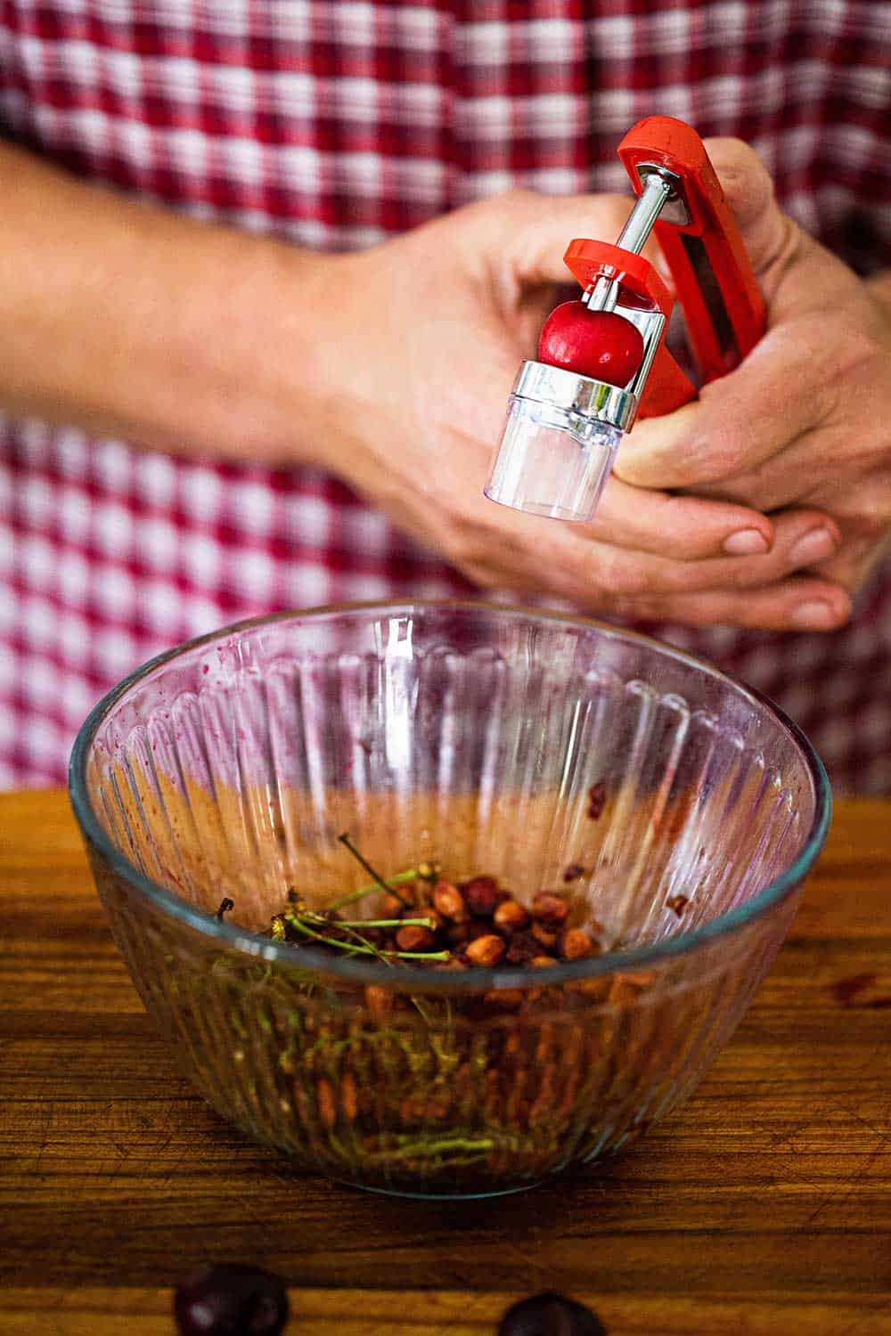 A person holding a cherry pitter with a cherry in it over a glass bowl filled with cherry pits.