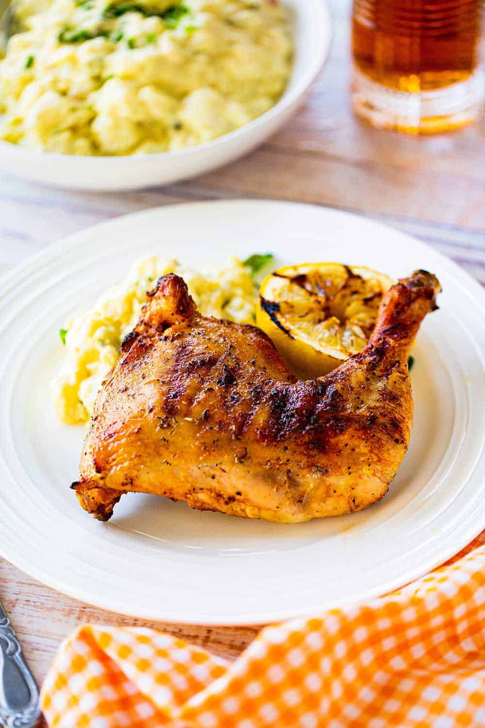A grilled chicken leg quarter sitting on a plate next to a grilled lemon half and potato salad.