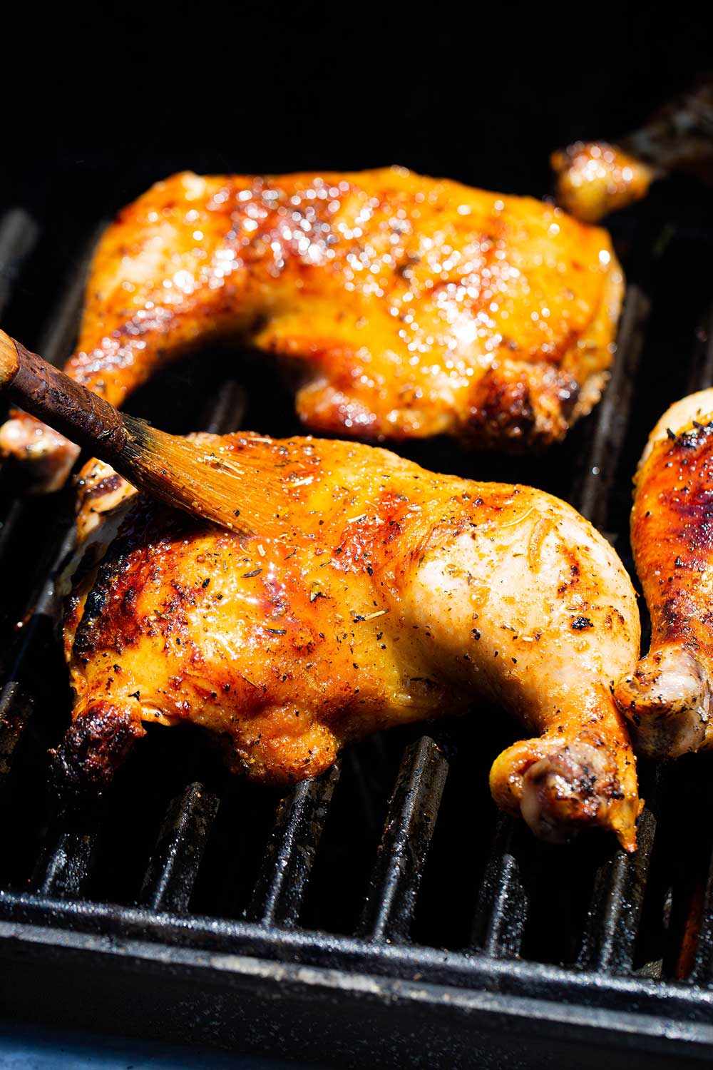 A chicken leg quarter on a gas grill being basted with a brush.