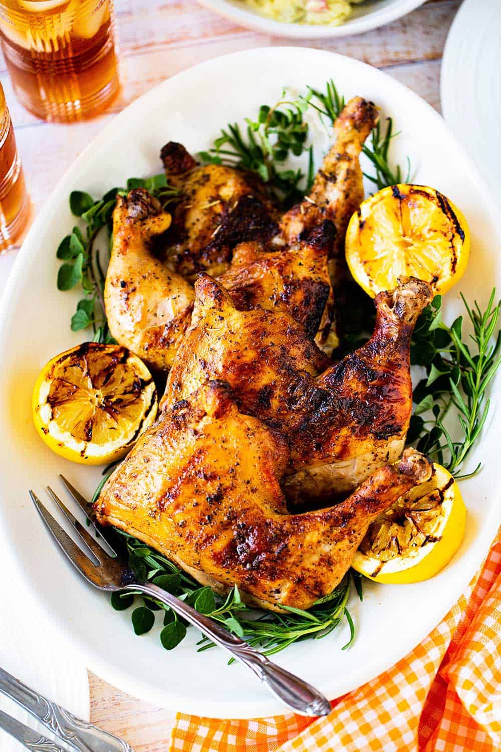 Three grilled chicken leg quarters on an oval platter surround by grilled lemon halves and fresh herbs.