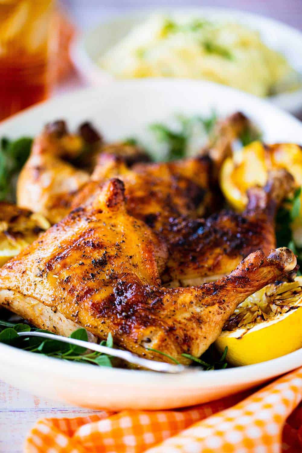 A grilled chicken leg quarter sitting on a platter next to grilled lemons and fresh herbs.