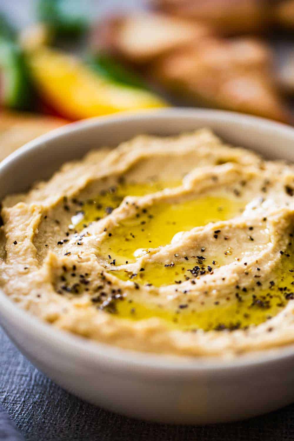 A bowl of homemade hummus with swirls of olive oil and black pepper on top all surrounded by cut bell peppers and pita chips. 