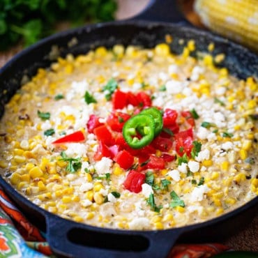 A cast-iron skillet filled with Mexican cream corn topped with crumbled queso fresco, chopped tomatoes, and chopped cilantro.