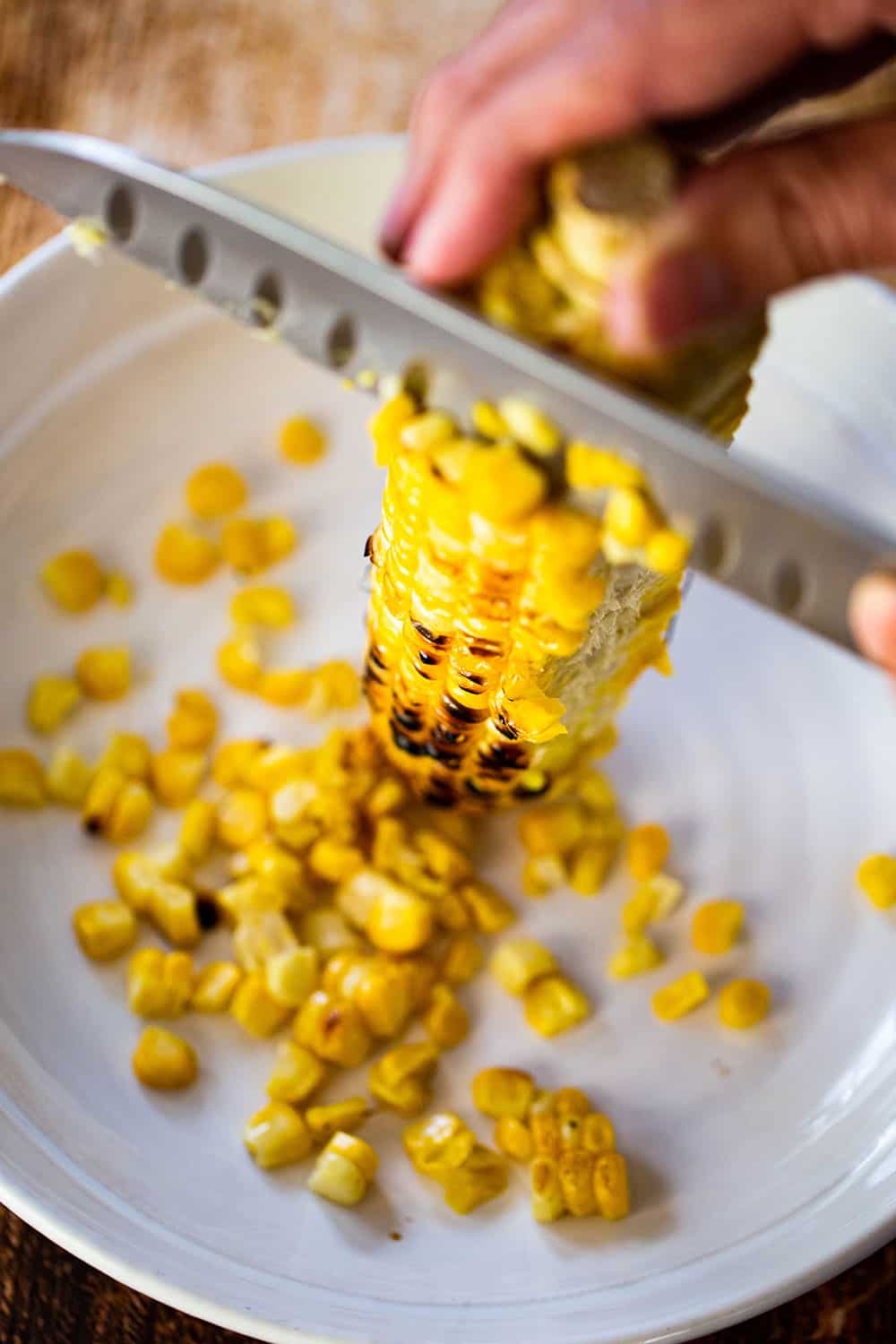 A person using a chef's knife to cut grilled corn kernels from an ear of a corn.