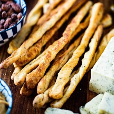 A pile of baked grissini, also known as Italian breadsticks, on a board next to a block of cheese that has been sliced and a bowl of spiced nuts.