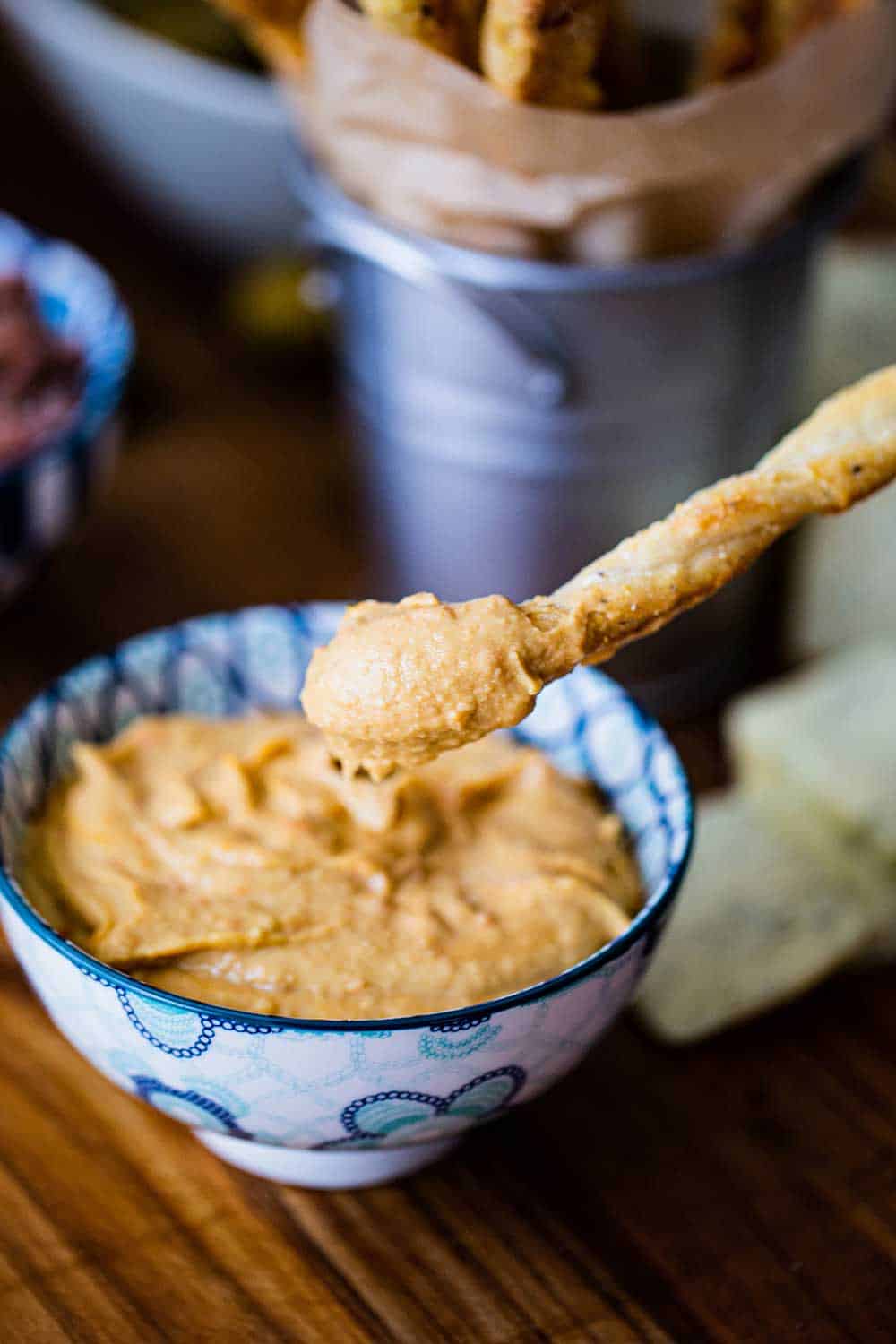 A breadstick that has been plunged into a bowl of hummus and being held just over the top of the dip. 