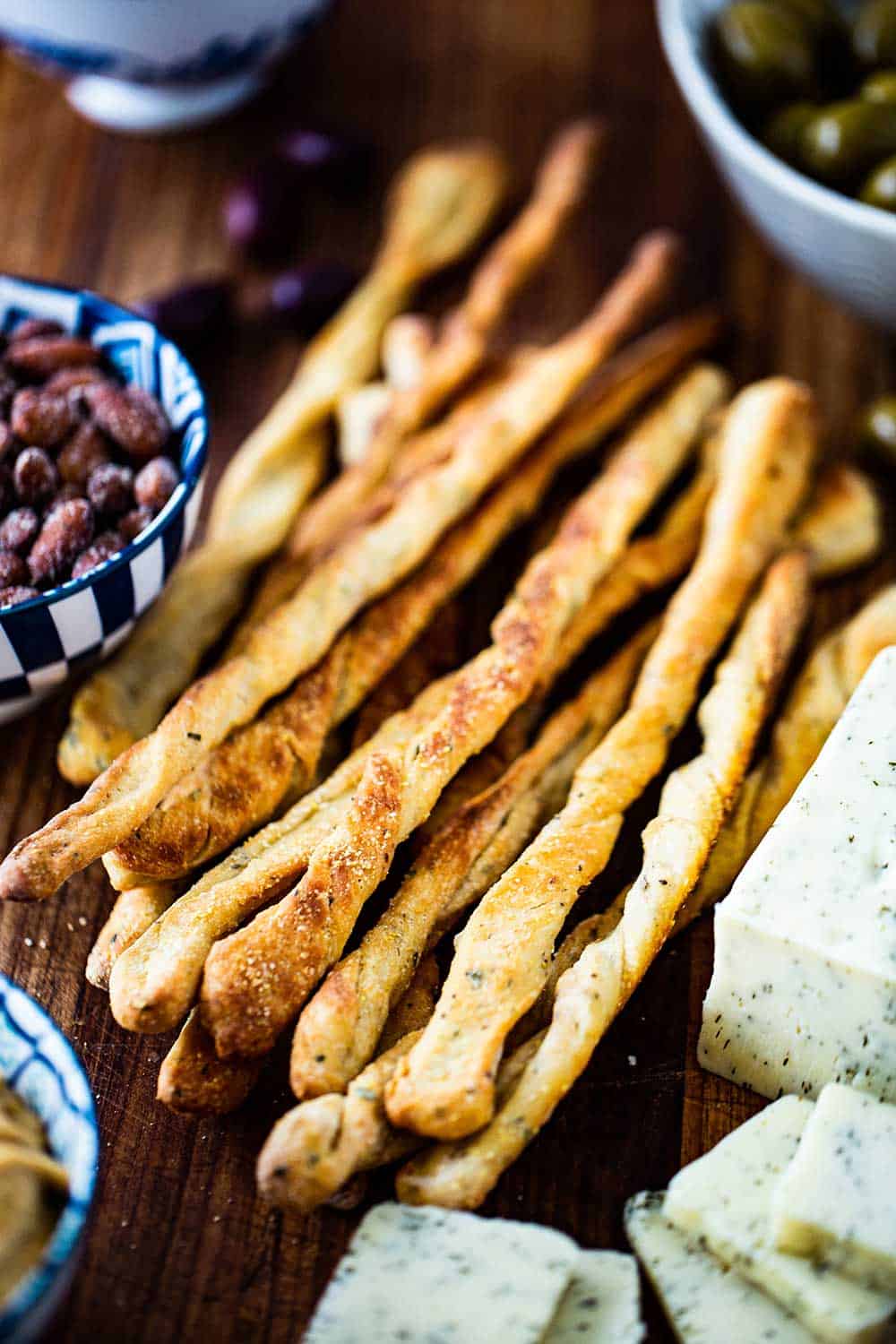 A pile of baked grissini, also known as Italian breadsticks, on a board next to a block of cheese that has been sliced and a bowl of spiced nuts. 