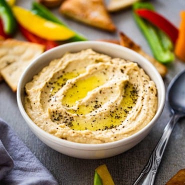 A bowl of homemade hummus with swirls of olive oil and black pepper on top all surrounded by cut bell peppers and pita chips.