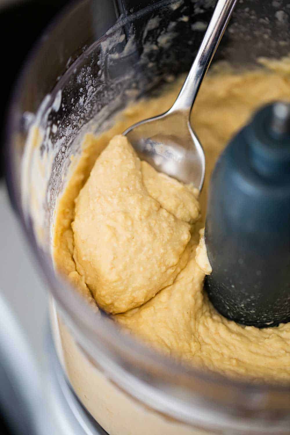 A spoon scooping up puréed hummus inside the bowl of a food processor. 