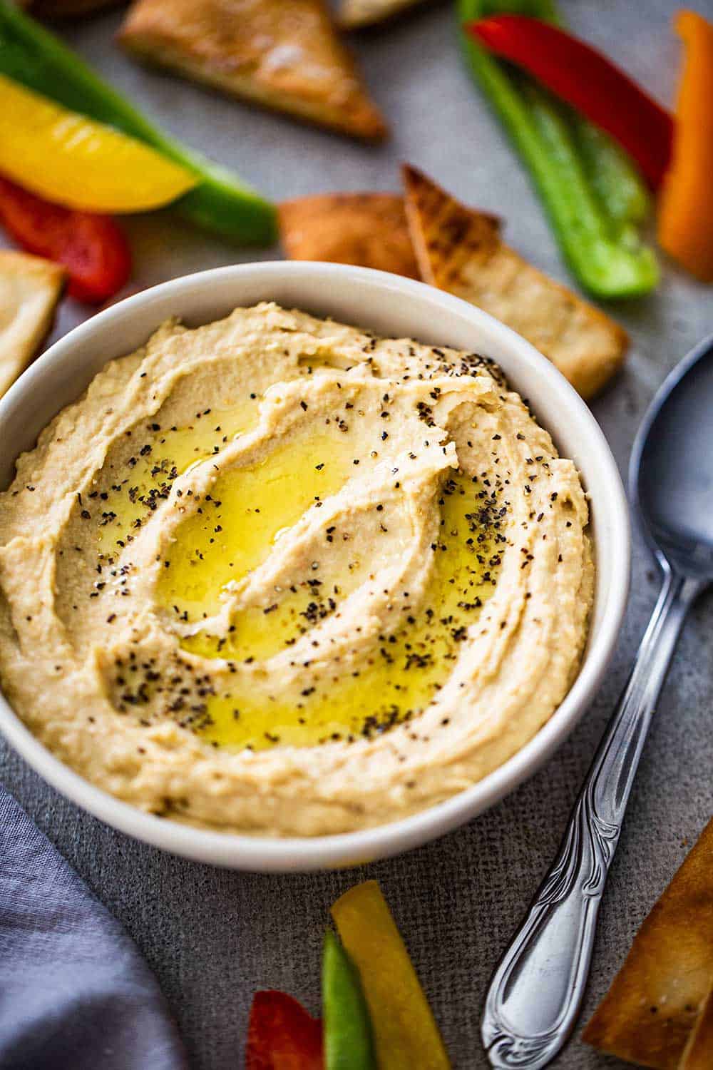 A bowl of homemade hummus with swirls of olive oil and black pepper on top all surrounded by cut bell peppers and pita chips. 