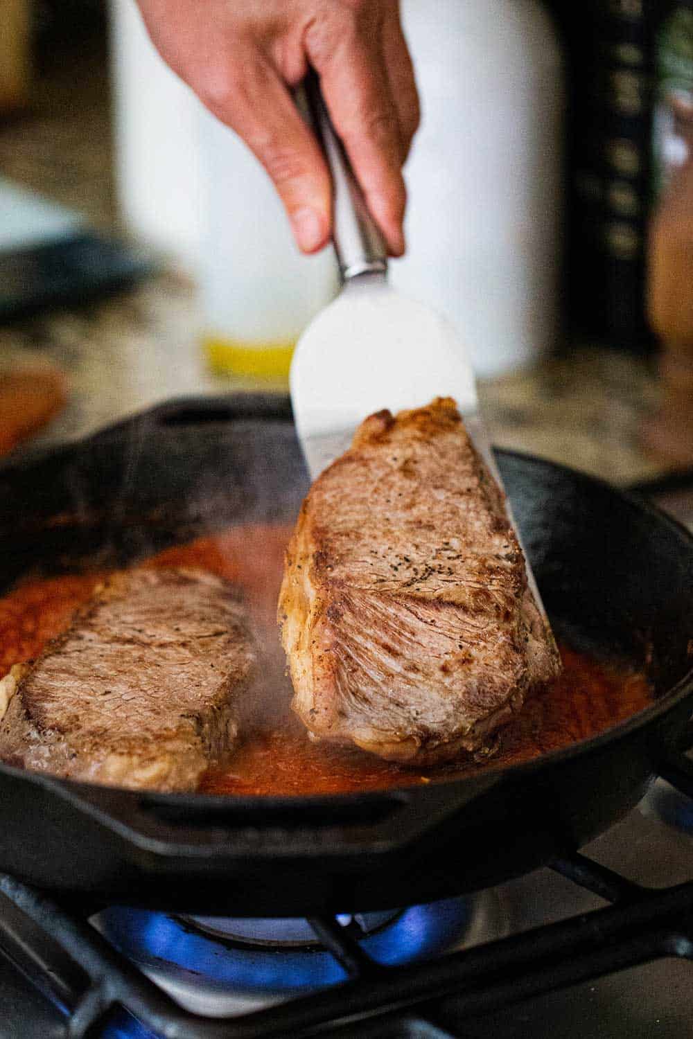 A person lowering a seared ribeye steak into a skillet filled with tomato sauce and another seared steak.