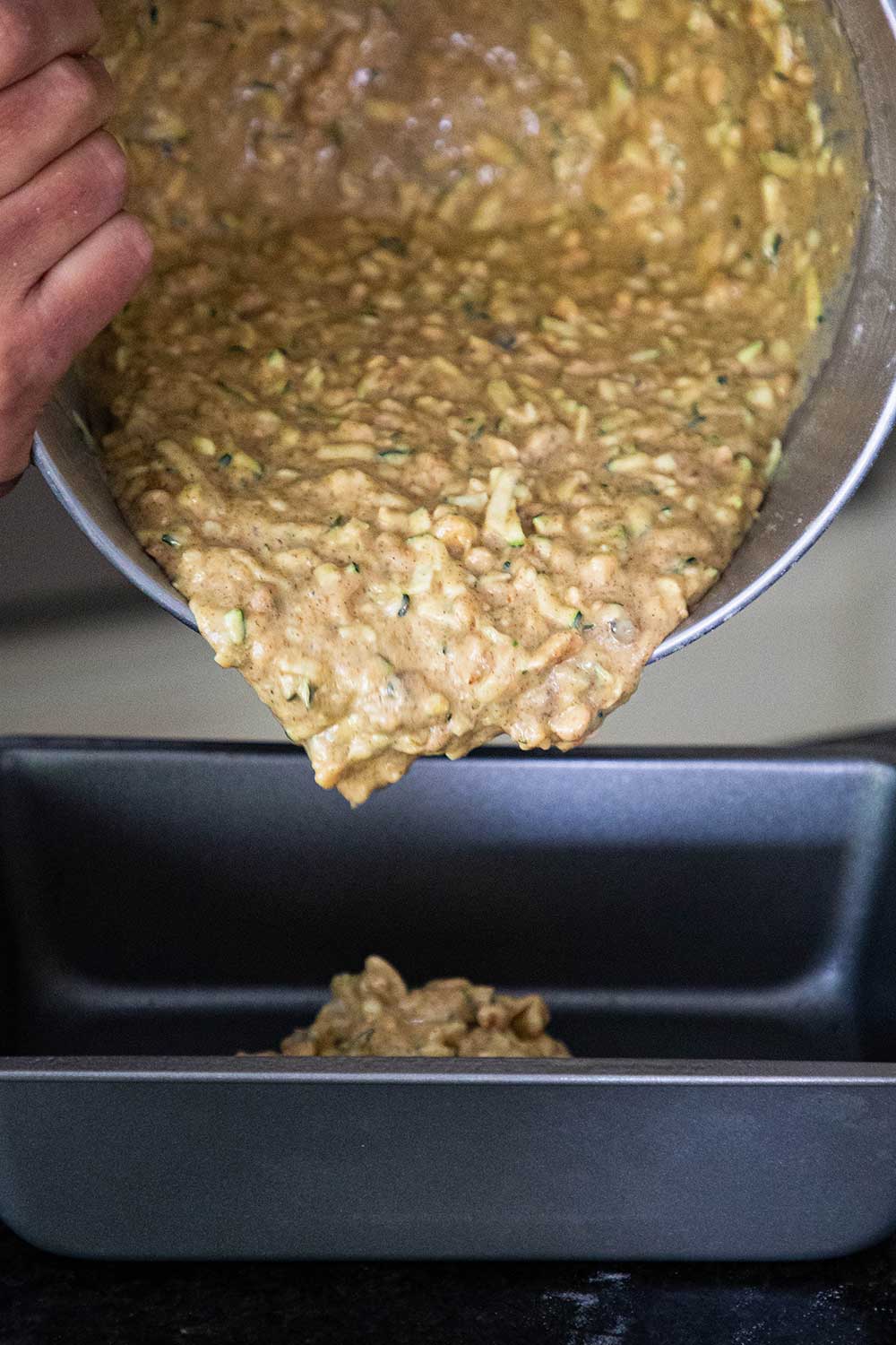 A person pouring zucchini batter from a mixing bowl into a metal loaf pan. 