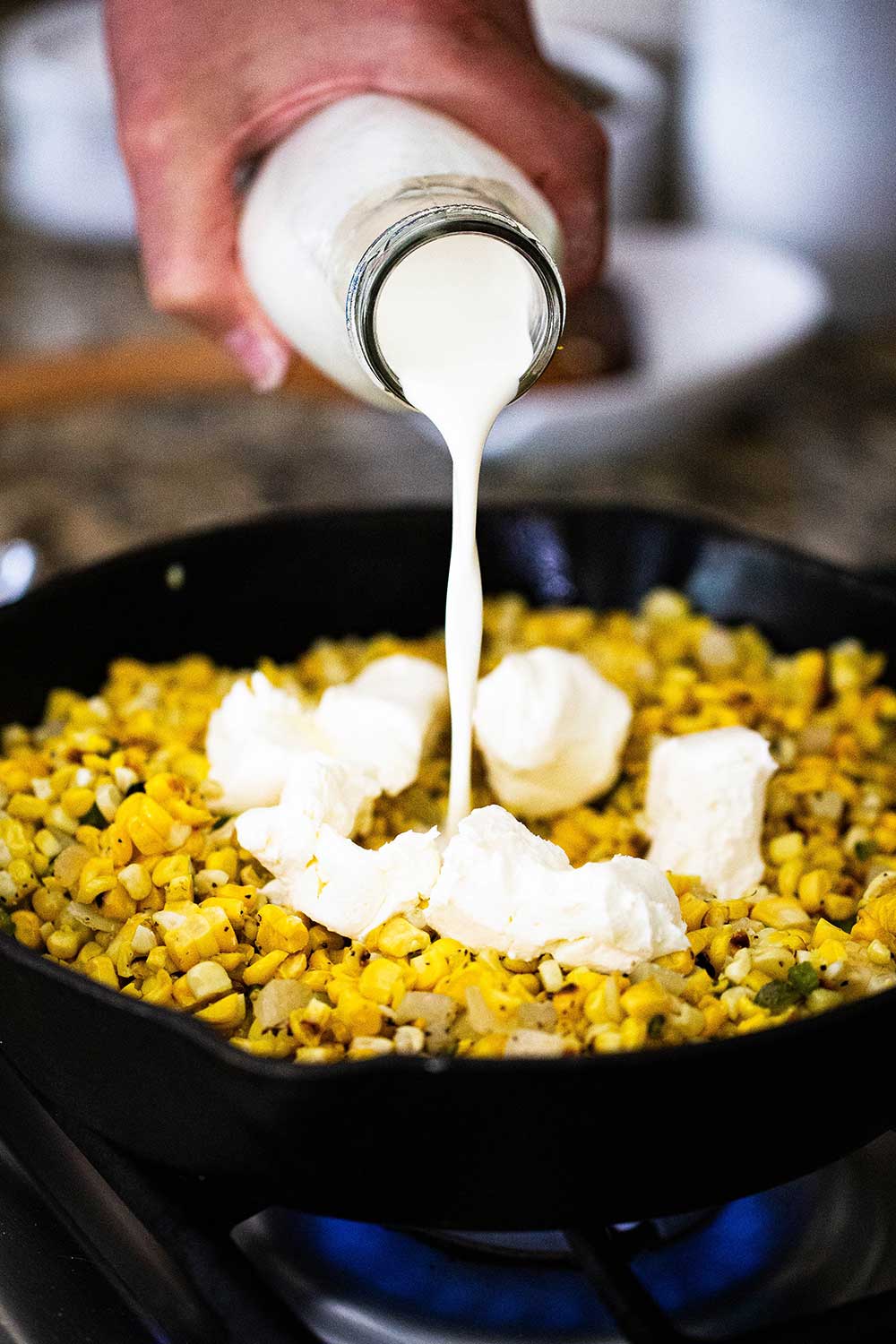 A person pouring heavy cream from a dairy bottle into a cast-iron skillet filled with grilled corn kernels, and chunks of cream cheese.