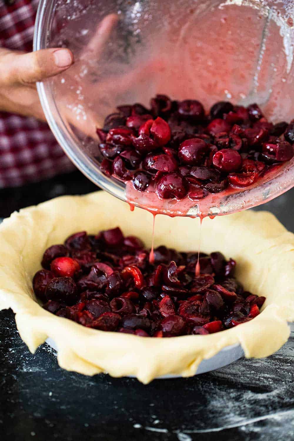 A person pouring cherry pie filling from a glass bowl into a pie dish that is lined with pie dough.