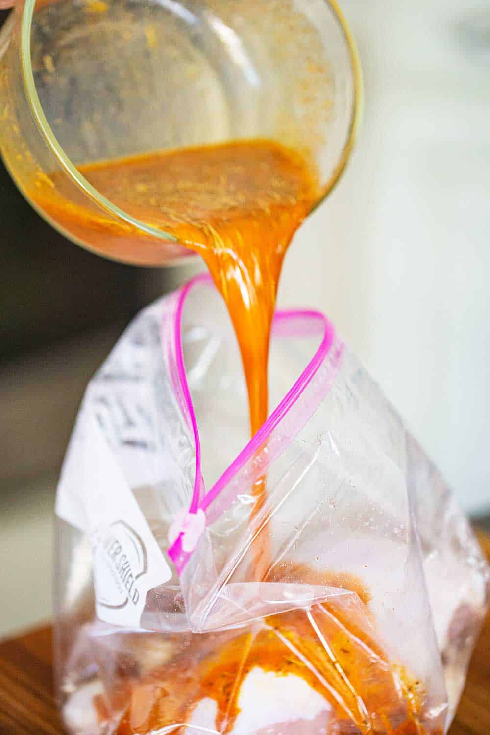An orange colored marinade being poured from a measuring cup into a plastic freezer baggie with chicken pieces in it.