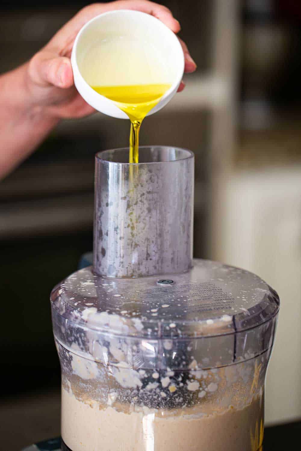 A person pouring extra-virgin olive oil from a small white bowl through the feeder of a food processor that is filled with puréed hummus. 