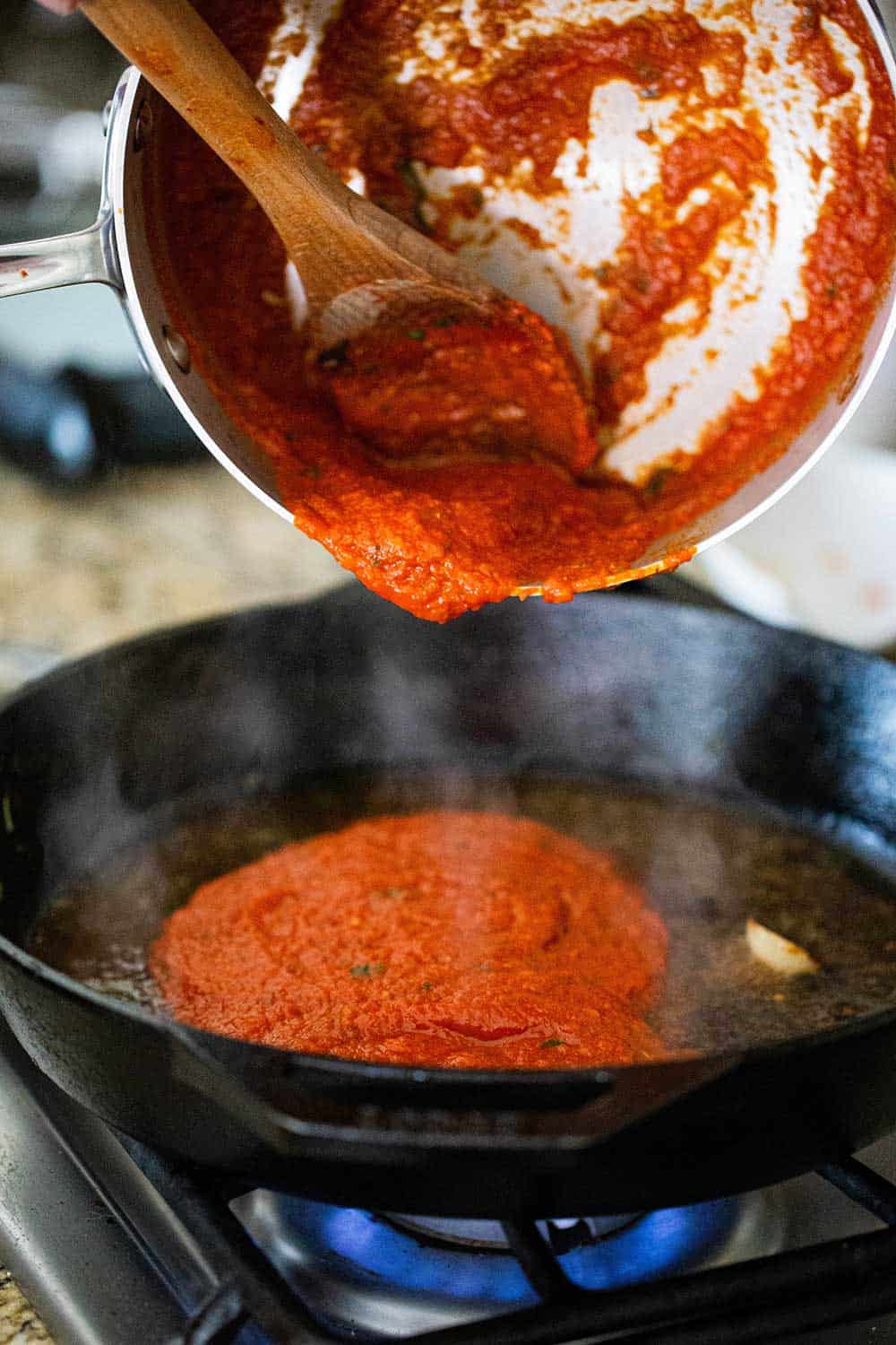 A medium-sized pot being held over a cast-iron skillet on a stove with tomato sauce transferring from the pot to the skillet.