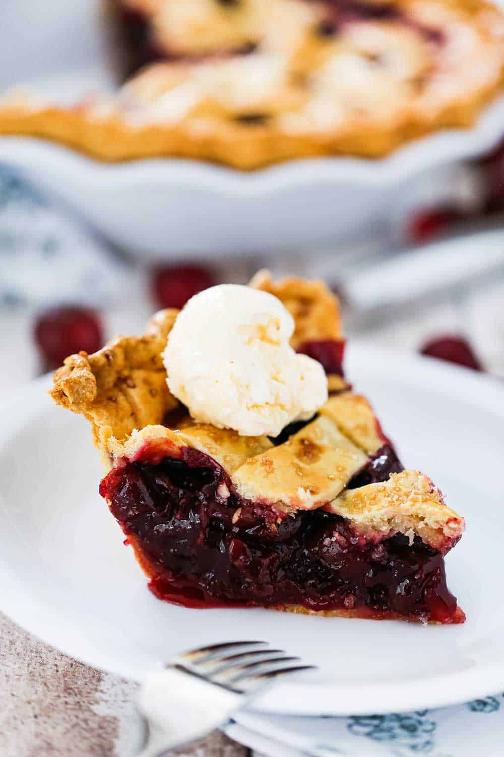 A single slice of cherry pie on a white dessert plate with a scoop of vanilla ice cream on top.