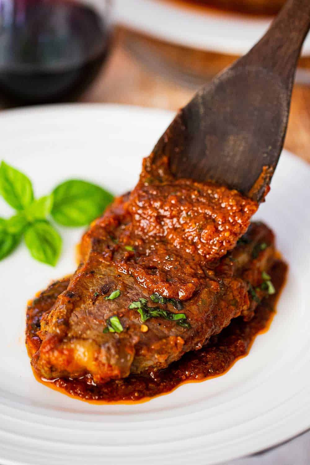 A wooden spoon transferring sauce onto steak pizzaiola on a white plate next to several basil leaves.