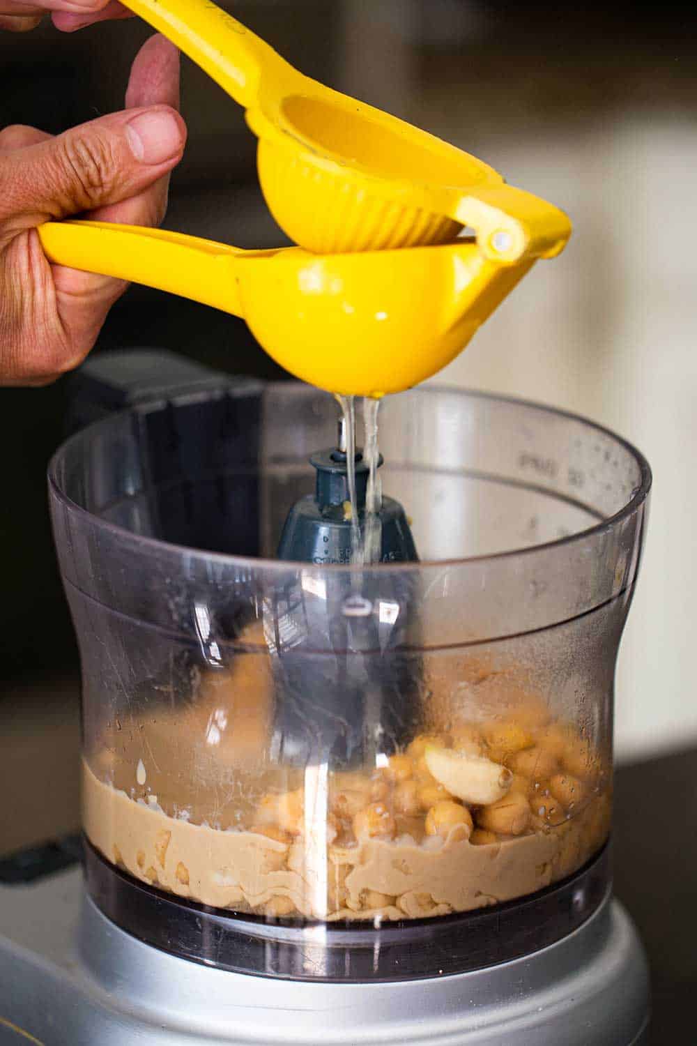 A person using a yellow citrus press to squeeze lemon juice from a lemon into a food processor filled with chickpeas and tahini for hummus. 