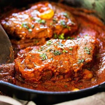 A cast-iron skillet filled with to seared steaks covered in a tomato sauce for steak pizzaiola.