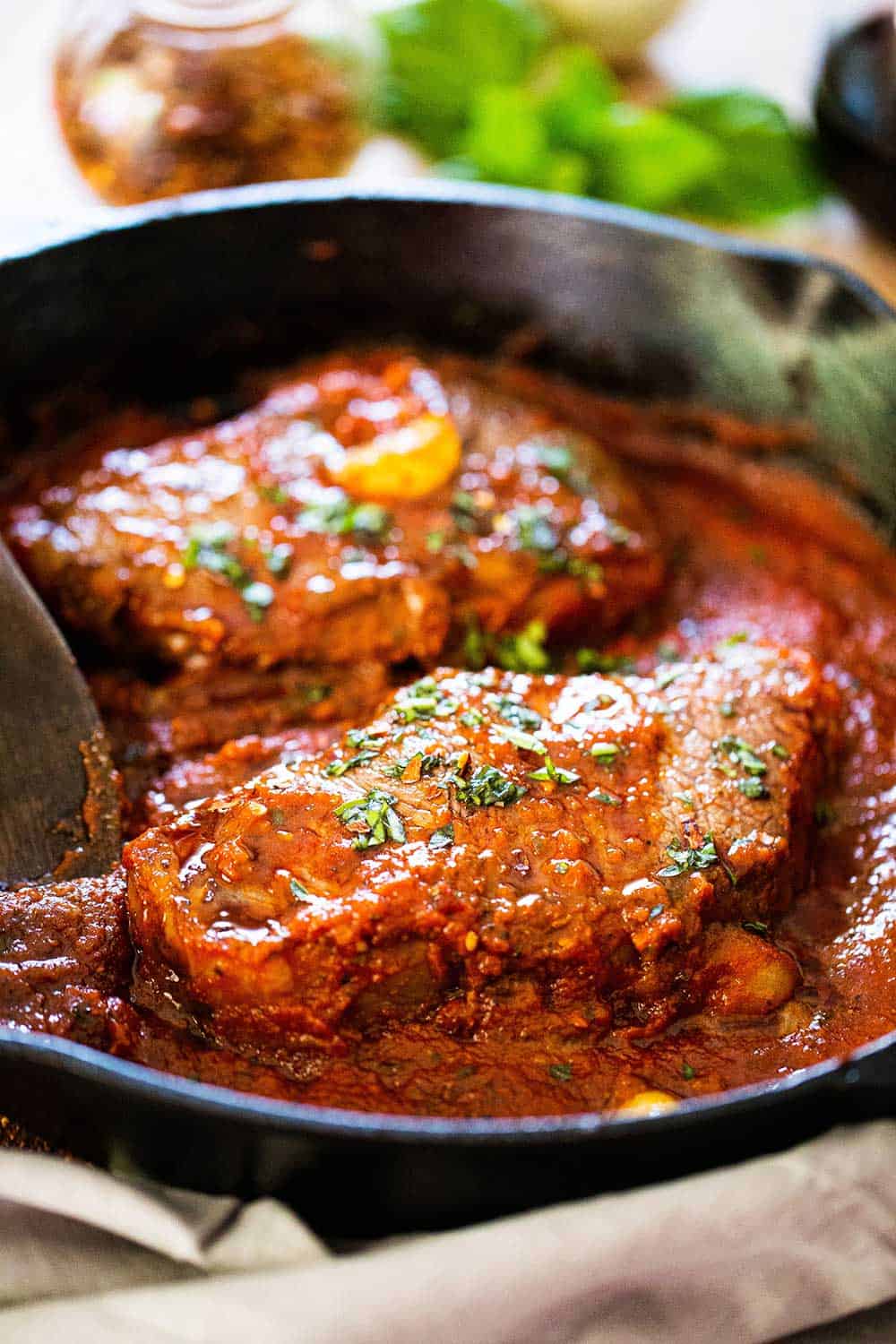 A cast-iron skillet filled with to seared steaks covered in a tomato sauce for steak pizzaiola.